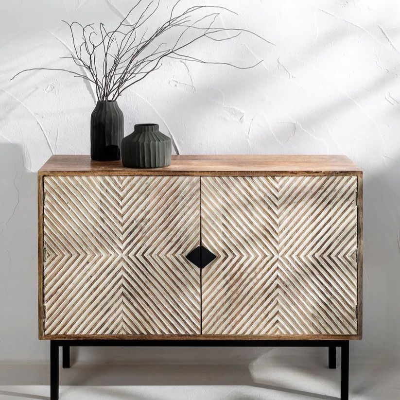 A carved wooden sideboard with a geometric pattern in an antique white finish, featuring a rectangular shape with a circular detail on the front, placed against a wall with decorative items on top.