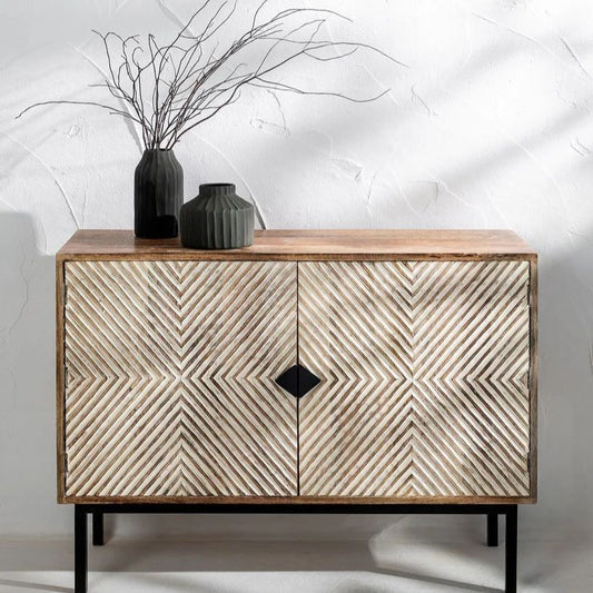 A carved wooden sideboard with a geometric pattern in an antique white finish, featuring a rectangular shape with a circular detail on the front, placed against a wall with decorative items on top.