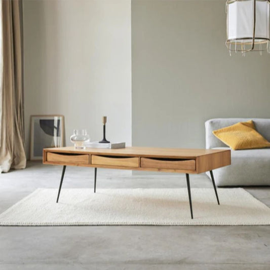 A rectangular solid wood center table with a Sheesham wood top and four foldable legs made of rust-proof metal, placed on a white rug in a minimalist living room setting with a sofa in the background and a lamp and a book on the table.