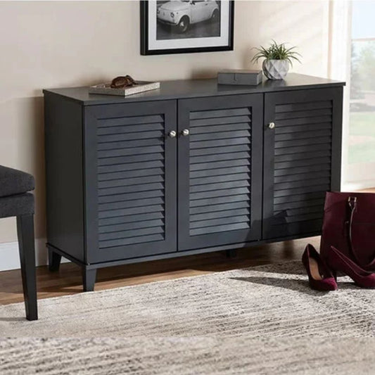 A gray shoe cabinet with three slatted doors and round knobs, placed in a home setting next to a chair and a plant, with a pair of shoes in front.